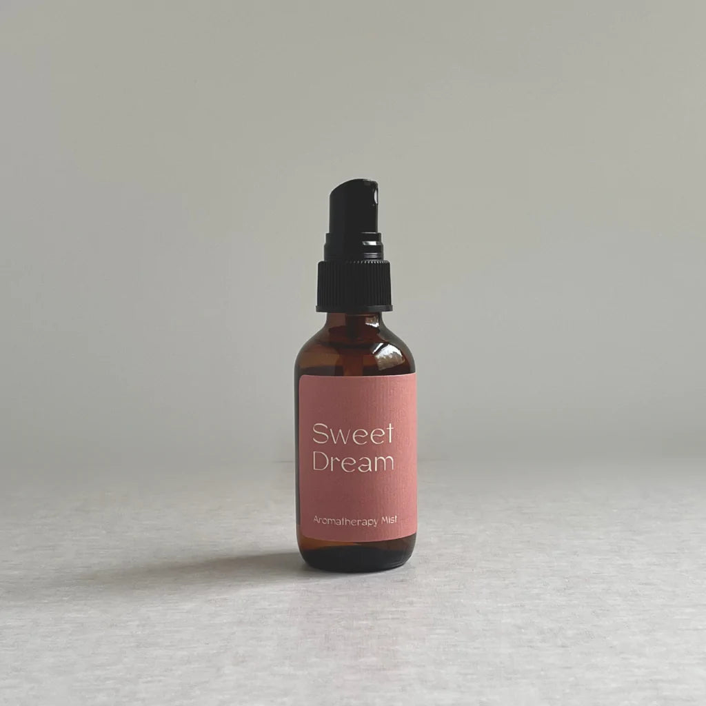 A small amber spray bottle with a black pump top sits on a light surface. Its pink label reads Sweet Dream Aromatherapy Mist by Species by the Thousands - Faire, crafted with organic essential oils for a calming citrus scent. The background is plain and neutral.