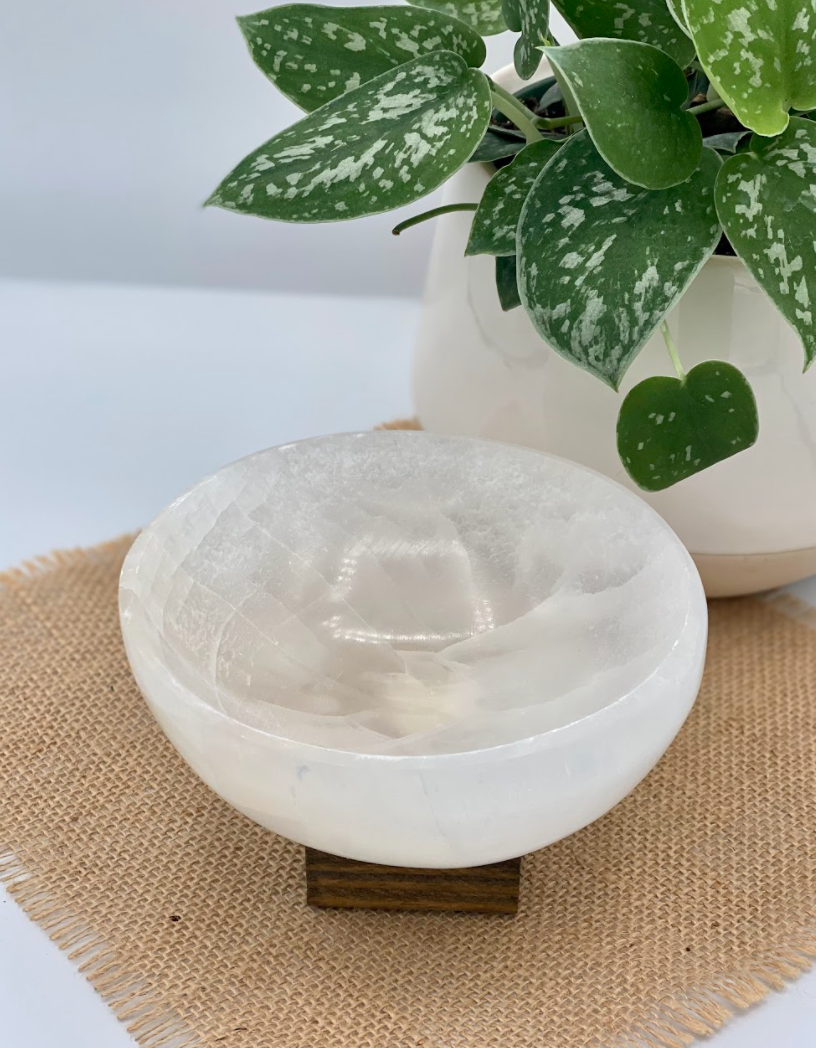 A Large Selenite Bowl by The Healing Hedge Witch sits on a small wooden stand atop a burlap mat, with a green potted plant partially visible in the background.