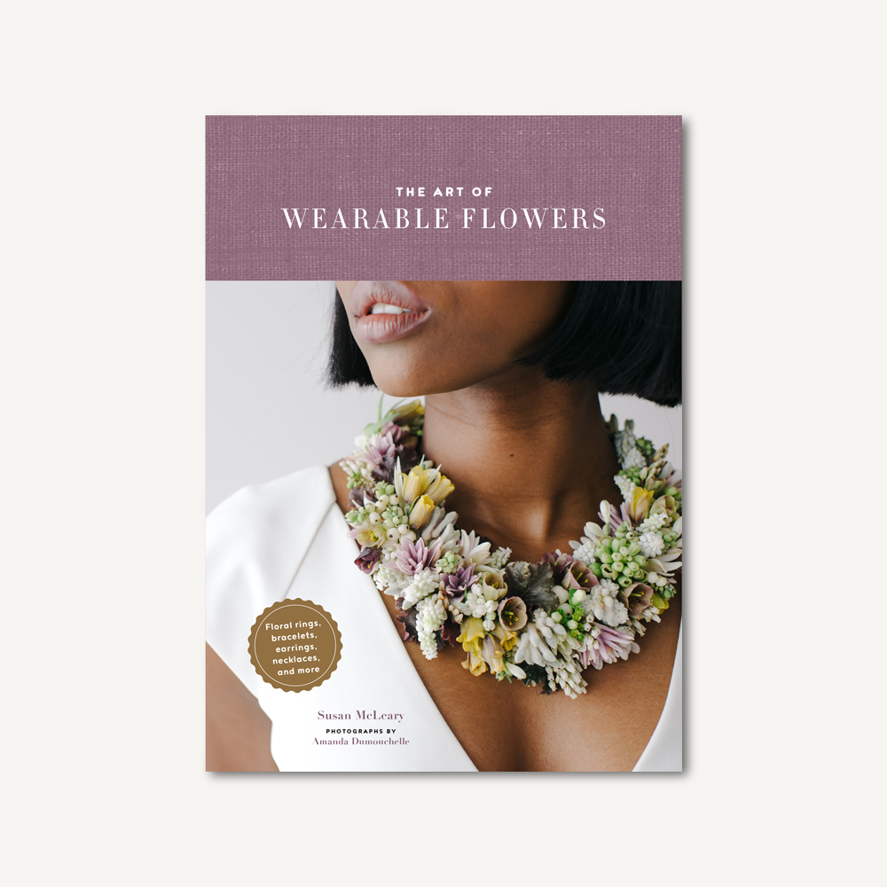 The cover of The Art of Wearable Flowers shows a woman adorned with an elaborate floral necklace against a light background, with the title displayed on a textured purple banner—ideal inspiration for flower arrangement lovers.