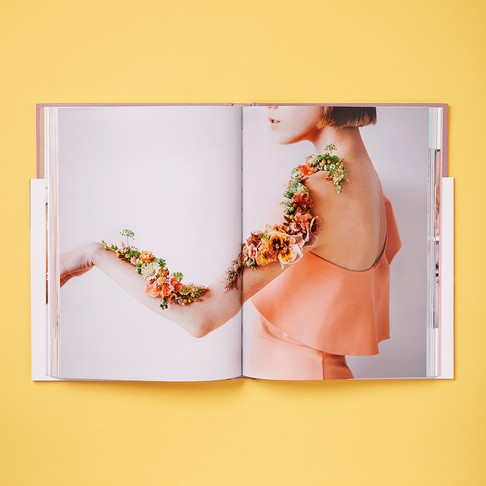 The Art of Wearable Flowers book is open to a photo of a woman in a peach dress with extended arm, displaying wearable flowers and greenery on a light background above a yellow surface.